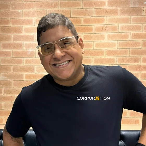 Integrante da equipe Corporaition sorrindo, de camiseta preta com a marca, diante de uma parede de tijolos